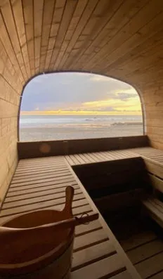 Inside a wooden sauna, looking out through an arched window at the sea at sunset